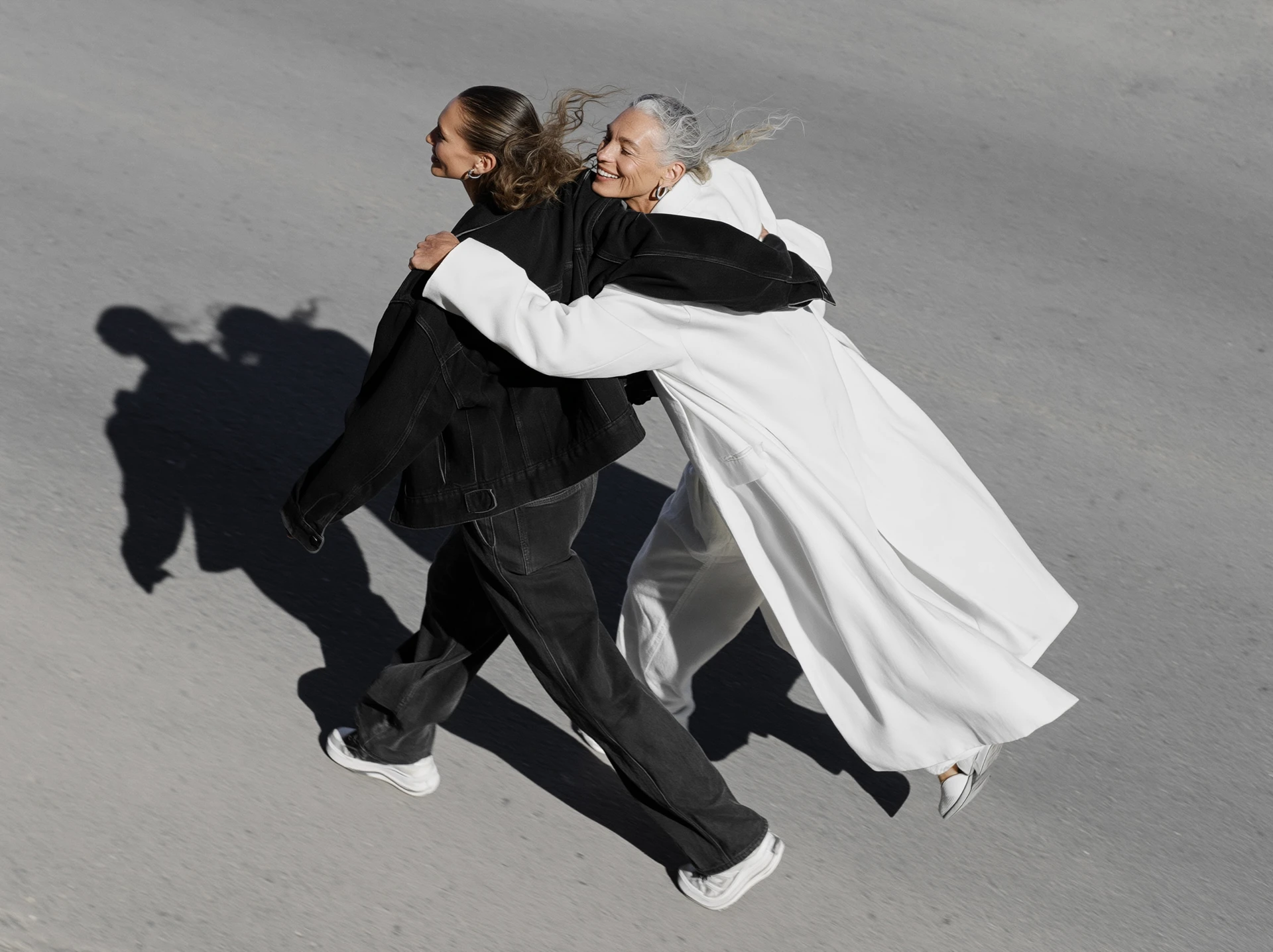 Two women walking arm in arm across an open street, one leaning in with a smile