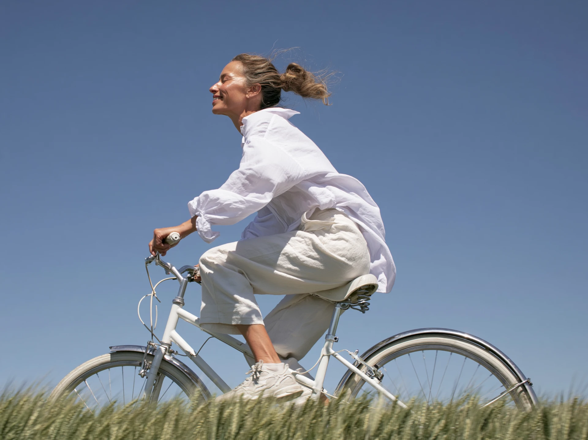 Woman riding a bicycle past a wheat field under a clear sky, eyes closed, enjoying the breeze