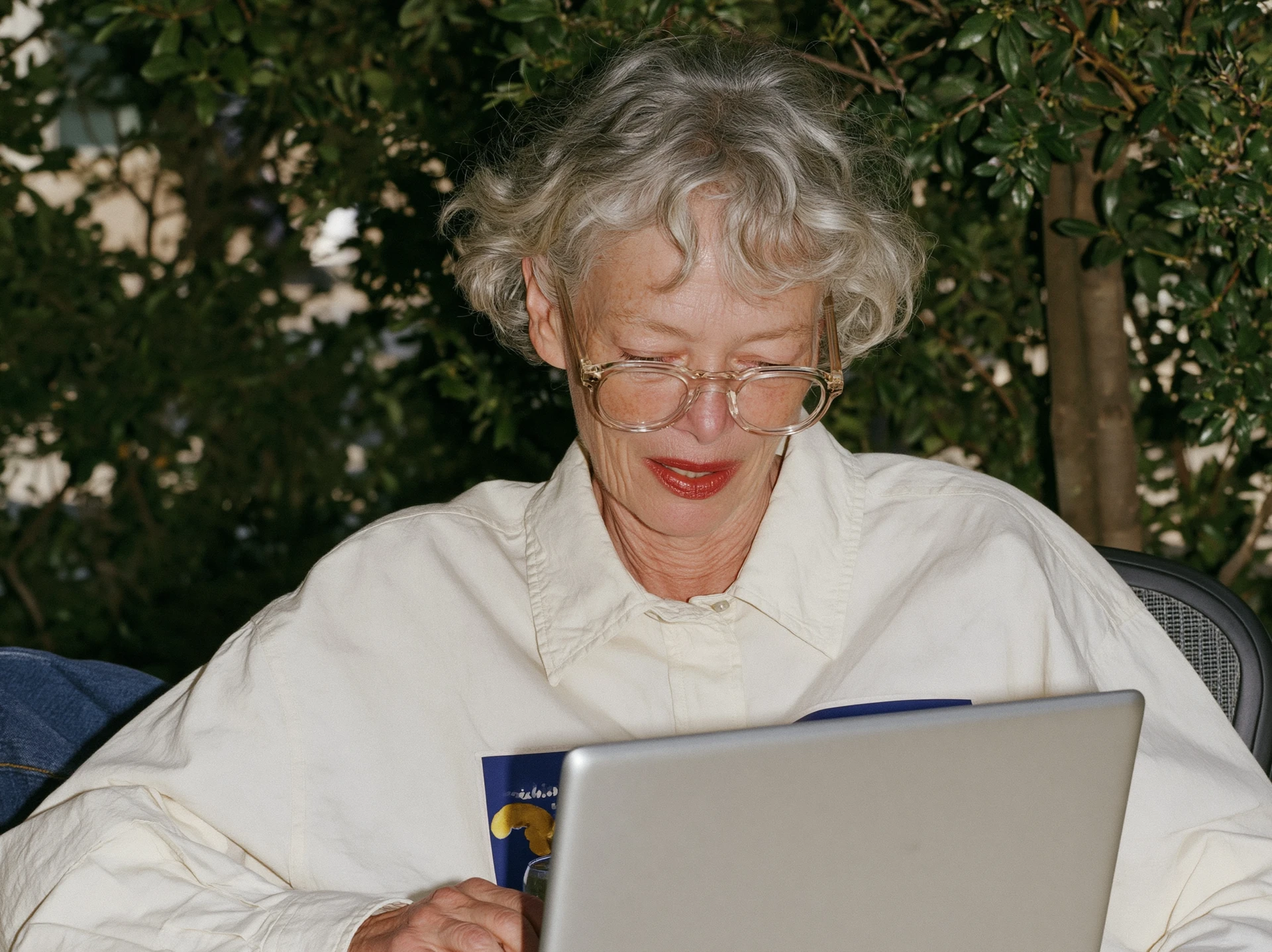 Woman with curly grey hair smiling at a laptop in a sunlit garden